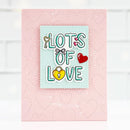 A pink handmade-style greeting card featuring a central layered white and light blue label with the words “LOTS OF LOVE” in teal outlined letters, decorated with small heart and key illustrations.