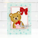 A handmade greeting card featuring a brown teddy bear holding a red heart, framed by a pink scalloped border with a red bow on a light blue background with white hearts and the word 'hugs' in script.