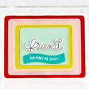 A handmade layered card featuring concentric rectangular paper borders in red, orange, yellow, peach, and pink, with a central mint green panel. The card has a die-cut white script word 'friend' and a teal banner reading 'YOU MAKE ME SMILE'.