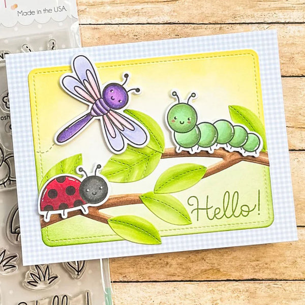 A handmade greeting card featuring cute cartoon bugs and leaves, displayed on a wooden surface next to a clear stamp sheet. The card has a white gingham border and a central yellow-green panel with a purple dragonfly, green caterpillar, red ladybug, and the word 'Hello!'.