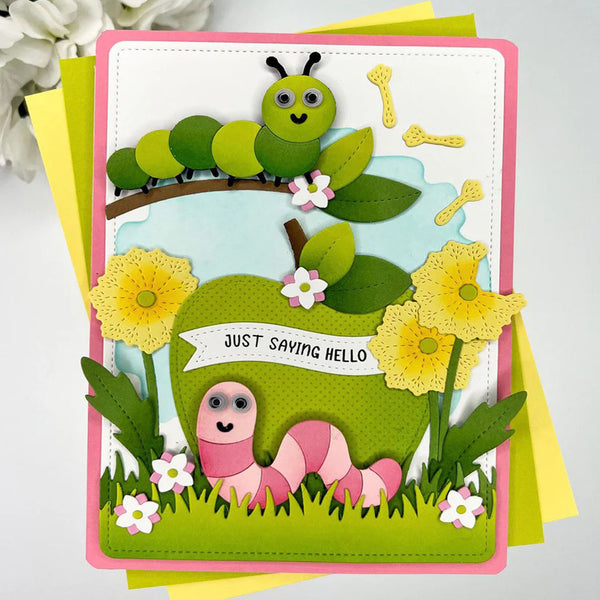 A handmade children's greeting card featuring layered paper crafts of two cute cartoon caterpillars, one green and one pink, along with yellow flowers, grass, and a banner that reads 'JUST SAVING HELLO'. The card is displayed on top of yellow and green envelopes against a light gray background with soft white flowers in the corner.