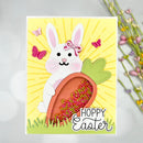 A handmade Easter greeting card featuring a white bunny holding a carrot filled with pink and green confetti, set against a yellow sunburst background with butterflies and the text 'Hoppy Easter'. The card is displayed on a light gray surface with blurred purple flowers in the background.