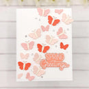 A handmade spring greeting card with layered paper butterflies in pink, peach, and coral orange, embellished with small clear rhinestones and a die-cut 'SPRING Greetings' label, set against a white wood plank background.