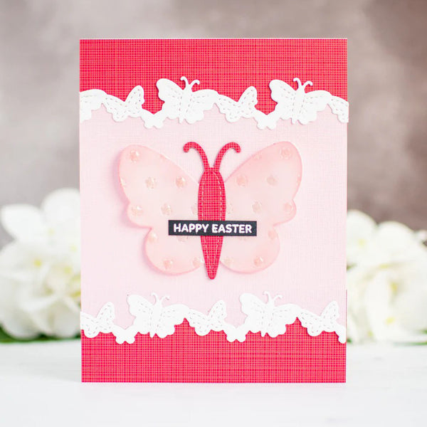 A handmade Easter greeting card featuring a pink and red butterfly design, white lace-like borders, and a black banner reading 'HAPPY EASTER', photographed against a softly blurred background with white flowers.