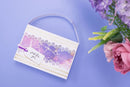 A white rectangular gift bag or clutch with a translucent handle, decorated with a purple and pink marbled pattern, white lace-like borders, and a small hanging tag that reads 'just for you'. It is photographed on a light lavender background with pink and purple flowers at the right edge.
