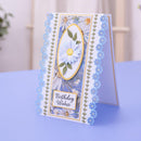 A handmade birthday card with a floral design, featuring a central white daisy in an oval frame, blue and white flower borders, and a 'Birthday Wishes' label, displayed on a light blue surface.
