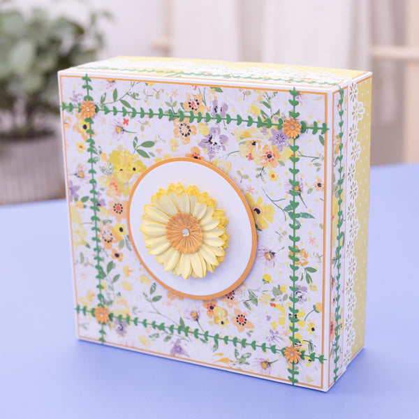 A decorative square gift box with a white background and a colorful floral print, featuring a large yellow flower applique on the front, set on a light blue surface.