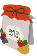 A decorative gift tag or card shaped like a jam jar, with a red lid, brown ribbon bow, and the phrase 'JAM-PACKED with love' on a white label. Fruit illustrations including strawberries, blueberries, raspberries, green leaves, and a lemon slice decorate the front, shown on a plain white background.