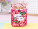 A decorative pink jar-shaped ornament or gift tag with a strawberry pattern, a gingham lid, twine bow, and a central label reading “JAM-PACKED with love,” photographed on a pastel surface with a blurred background.
