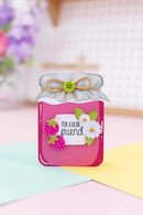A handmade greeting card shaped like a pink glass jar filled with jam, featuring a white lid with a tan ribbon bow and a green button. The front is decorated with a white daisy, a pink strawberry, green leaves, and a white tag that reads 'FOR A DEAR friend'.