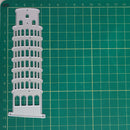 A white plastic or metal laser-cut template or stencil shaped like the Leaning Tower of Pisa, placed on a green cutting mat with grid lines.