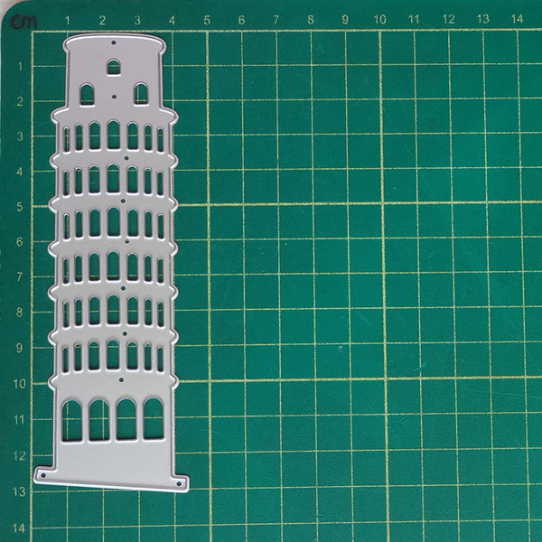 A white plastic or metal laser-cut template or stencil shaped like the Leaning Tower of Pisa, placed on a green cutting mat with grid lines.
