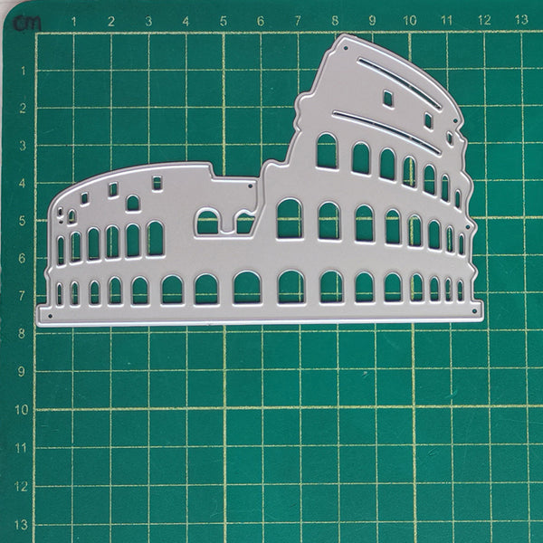 A silver metal die-cut stencil shaped like the Colosseum, displayed on a green cutting mat with grid lines.