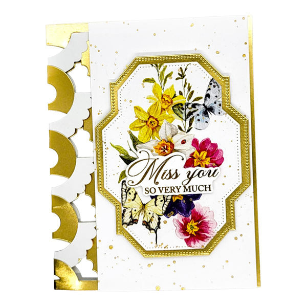 A handmade greeting card with a white background, gold foil accents, and a central octagonal label featuring colorful watercolor-style flowers and butterflies. The label reads 'Miss you SO VERY MUCH' in elegant script and serif text.