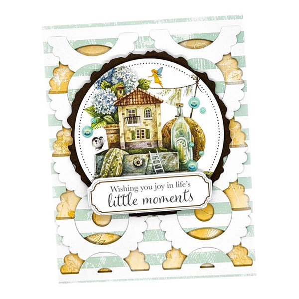 A handmade layered greeting card with a scalloped white outer border, a central circular illustration of a house and bottle, and a banner reading 'Wishing you joy in life's little moments'.