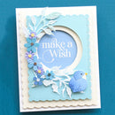 A handmade layered greeting card with a scalloped cream border, a light blue inner frame, and a central oval cutout containing the words 'make a Wish'. The card is decorated with white and blue paper leaves, blue and purple flowers, and a small blue bird, set against a solid teal background.