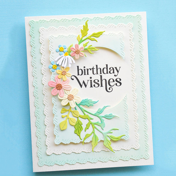 A handmade layered greeting card with scalloped white and pale mint borders, featuring a floral arrangement in pastel pink, yellow, blue, and green, with the words 'birthday wishes' in black script on a light blue background.