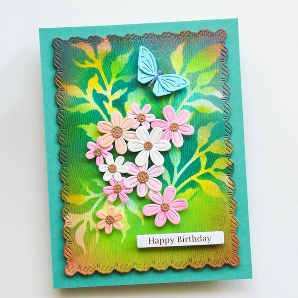 A handmade layered greeting card featuring a scalloped die-cut border, a green and yellow leafy background, pink and white daisy embellishments, a light blue butterfly, and a small white banner reading 'Happy Birthday'.