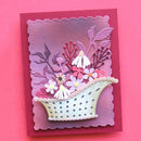 A handmade layered card featuring a basket filled with white, pink, and purple flowers, set against a purple gradient background and mounted on a dark red cardstock base, photographed on a solid pink background.