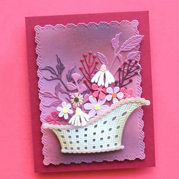 A handmade layered card featuring a basket filled with white, pink, and purple flowers, set against a purple gradient background and mounted on a dark red cardstock base, photographed on a solid pink background.