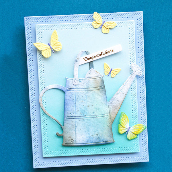 A square layered card featuring a distressed silver watering can illustration on a light blue background, decorated with yellow and lavender butterflies and a small 'Congratulations' banner, set against a solid blue backdrop.