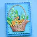 A square handmade paper craft card featuring a woven brown basket filled with yellow, orange, and pink flowers, green leaves, and white berries, set against a layered blue background with a scalloped border.