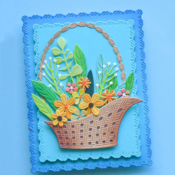 A square handmade paper craft card featuring a woven brown basket filled with yellow, orange, and pink flowers, green leaves, and white berries, set against a layered blue background with a scalloped border.
