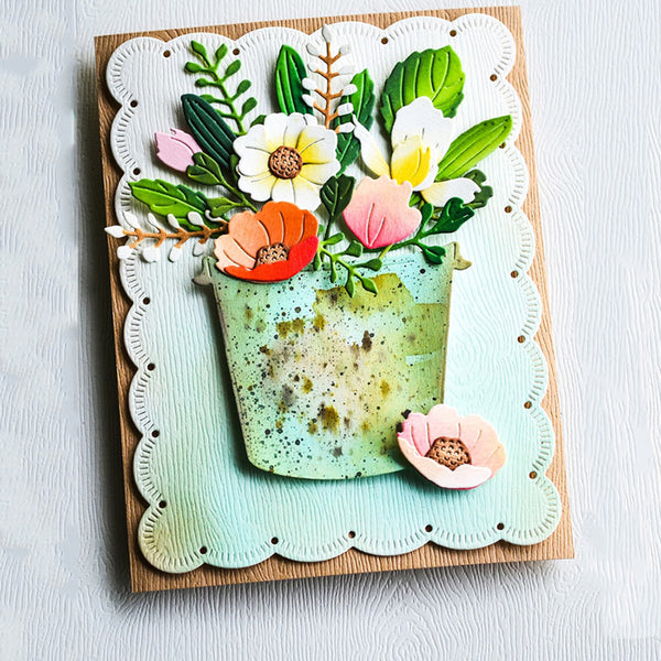 A handmade layered card featuring a 3D floral arrangement in a speckled green pot, set on a scalloped white die-cut border over a light wood background.