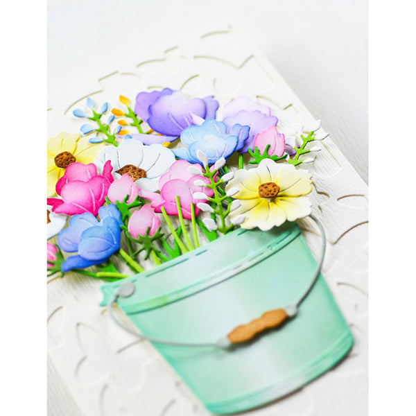 A decorative artificial flower arrangement in a small mint green bucket with a metal handle and wooden accent, photographed on a white textured background.