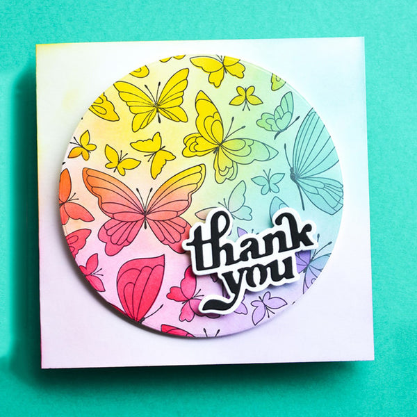 A square white card featuring a circular sticker or decal with a pastel rainbow gradient background, decorated with colorful hand-drawn butterflies and the words 'thank you' in black script with a white outline, set against a solid teal background.