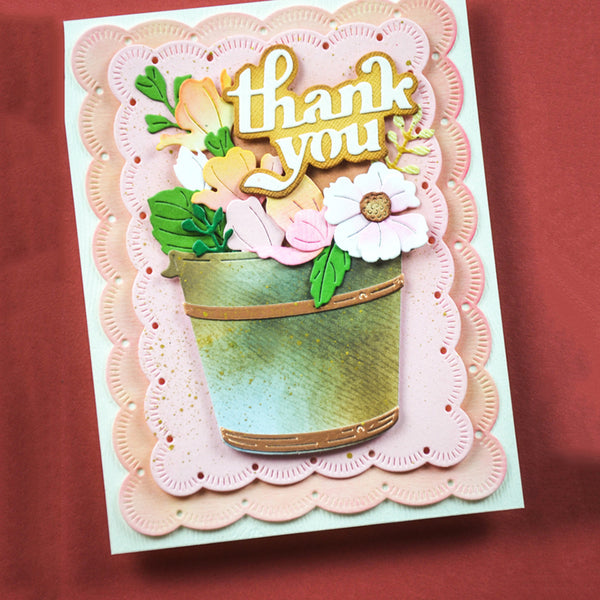 A handmade layered greeting card featuring a scalloped pink and peach border, a green and gold watering can, and layered paper flowers and leaves with the words 'thank you' in gold foil lettering. The card is photographed flat on a textured dark red background.