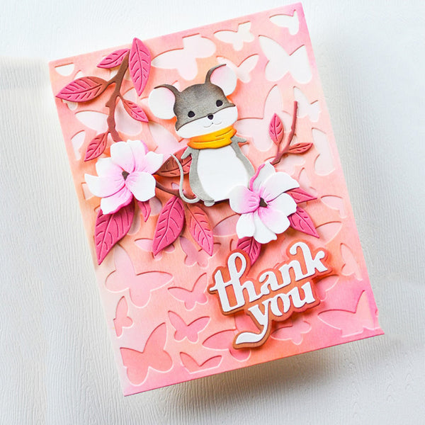 A handmade layered greeting card featuring a cute mouse character wearing a yellow scarf, surrounded by pink and white flowers, butterfly cutouts, and a 'thank you' message on a pink watercolor background.