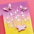 A handmade layered greeting card featuring white lily-of-the-valley flowers, pink butterflies, and a pink 'thank you' message on a pink-to-yellow gradient cardstock background.