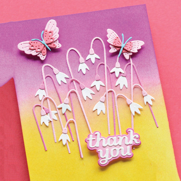 A handmade layered greeting card featuring white lily-of-the-valley flowers, pink butterflies, and a pink 'thank you' message on a pink-to-yellow gradient cardstock background.