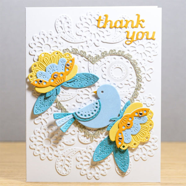 A handmade layered greeting card featuring a white embossed lace background, a light blue bird, two yellow and teal butterfly embellishments, and gold foil 'thank you' lettering at the top right.