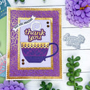 A handmade greeting card featuring a purple coffee cup and the words 'thank you' and 'YOU ARE AMAZING' in purple and gold, styled on a white wood background with decorative flowers and greenery.
