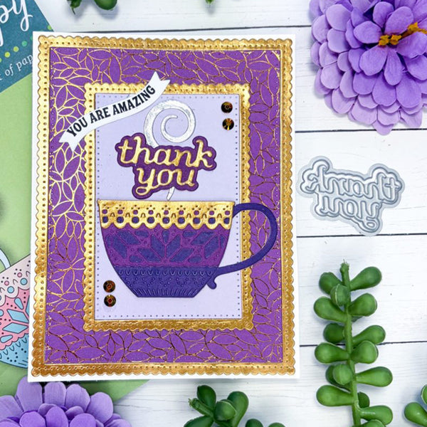 A handmade greeting card featuring a purple coffee cup and the words 'thank you' and 'YOU ARE AMAZING' in purple and gold, styled on a white wood background with decorative flowers and greenery.