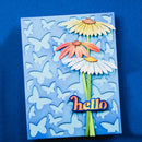 A handmade layered paper card featuring three colorful daisy flowers on a light blue background with cutout butterflies and the word 'hello' in orange script at the bottom.