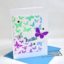 A handmade greeting card with a white base, featuring a layered butterfly design in green, blue, and purple on a white background. The word 'hello' is written in blue script on a small white label, and a purple butterfly is layered over it.