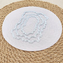 Three nested oval die-cut metal cutting mats with scalloped edges, displayed on a round white sheet over a woven jute placemat.