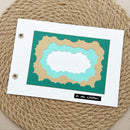 A handmade layered paper frame with scalloped edges in teal, light blue, and tan, mounted on a white card with two binder holes. The card is placed on a round woven jute placemat, with a small black label reading 'In der Welt' at the bottom.