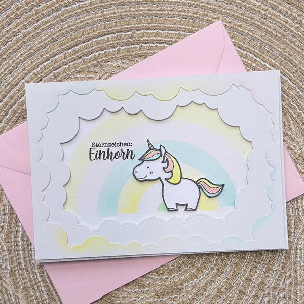 A handmade greeting card with a scalloped white border and a cute unicorn illustration on a pastel rainbow background, placed on top of a pink envelope over a woven straw surface.