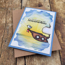 A handmade greeting card with a blue border and a colorful illustration of a seagull on a ship's deck, placed on top of a brown envelope over a rustic wooden surface. The card features German text that reads 'ICH SCHICKE DIR Sonnenschein'.