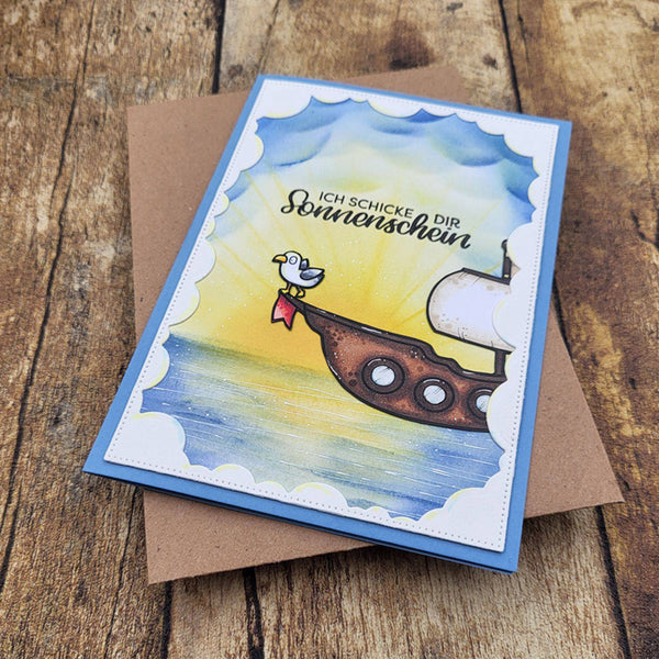 A handmade greeting card with a blue border and a colorful illustration of a seagull on a ship's deck, placed on top of a brown envelope over a rustic wooden surface. The card features German text that reads 'ICH SCHICKE DIR Sonnenschein'.
