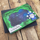 A handmade greeting card featuring a green scalloped border, a starry night sky background, a cartoon wizard character, and German text, displayed on a black cardstock backing over a rustic wooden surface.