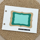 A handmade paper craft label with layered decorative frames in mint green, teal, and tan, mounted on a white cardstock sheet with two metal grommets on the left. The card is placed on a round woven jute placemat.