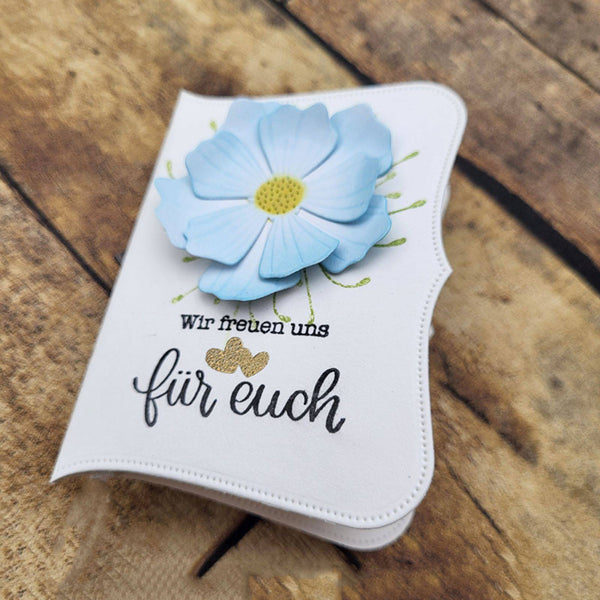 A handmade white card or small notebook with a large light blue flower on the front, black German text reading 'Wir freuen uns für euch', and gold glitter accents, photographed on a rustic wooden surface.
