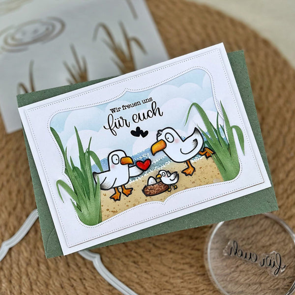 A handmade greeting card featuring a cute cartoon scene of three white seagulls, one adult and two chicks, standing on a sandy beach with green reeds, blue sky, and the German text 'Wir freuen uns für euch'. The card is layered over a sage green envelope and photographed on a textured tan surface with crafting tools nearby.