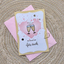 A handmade greeting card with a champagne toast illustration and German text, layered over a pink envelope on a woven straw mat.