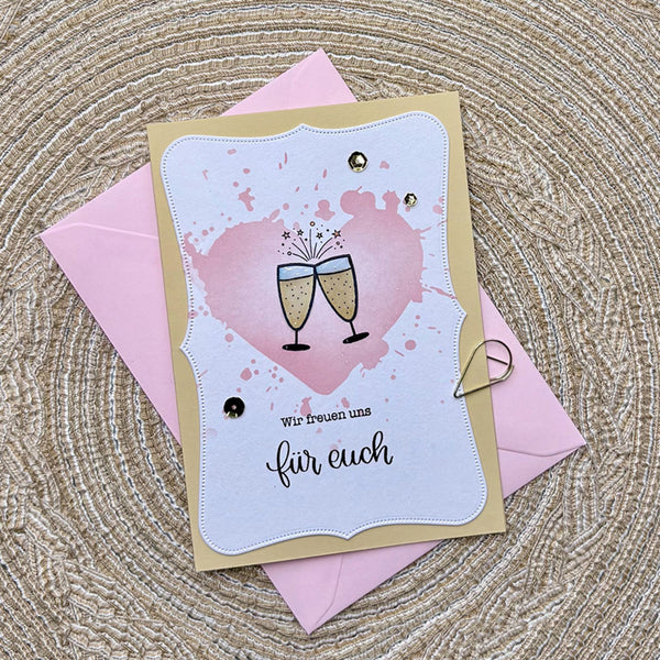 A handmade greeting card with a champagne toast illustration and German text, layered over a pink envelope on a woven straw mat.